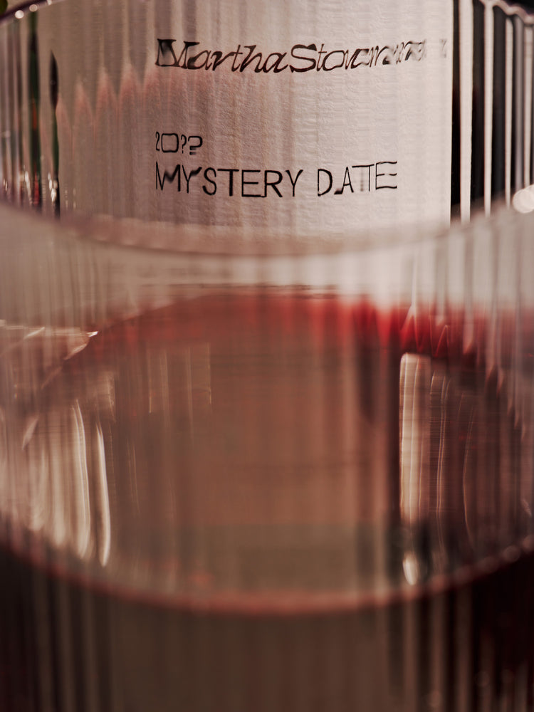 Close up of a blurred wine glass covering a "mystery date" wine label in the background.