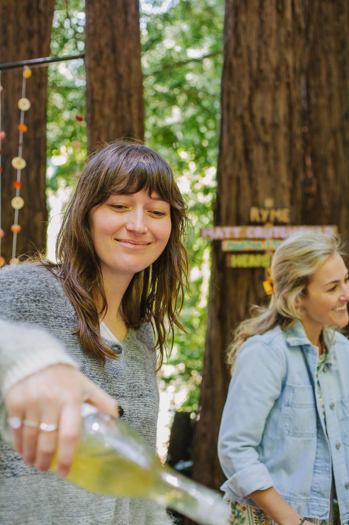 Two women enjoy a sunny day in the woods. One pours a drink smiling, while the other is laughing. Tall trees and a playful atmosphere surround them.