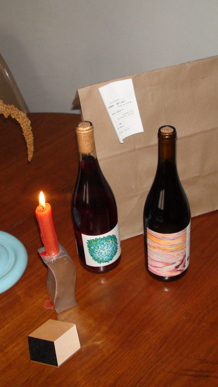 Two wine bottles on a wooden table with a lit candle and a take out bag.