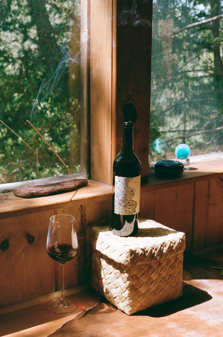 A wine bottle and glass sit on a woven basket near a window, with incense smoke rising and greenery visible outside.
