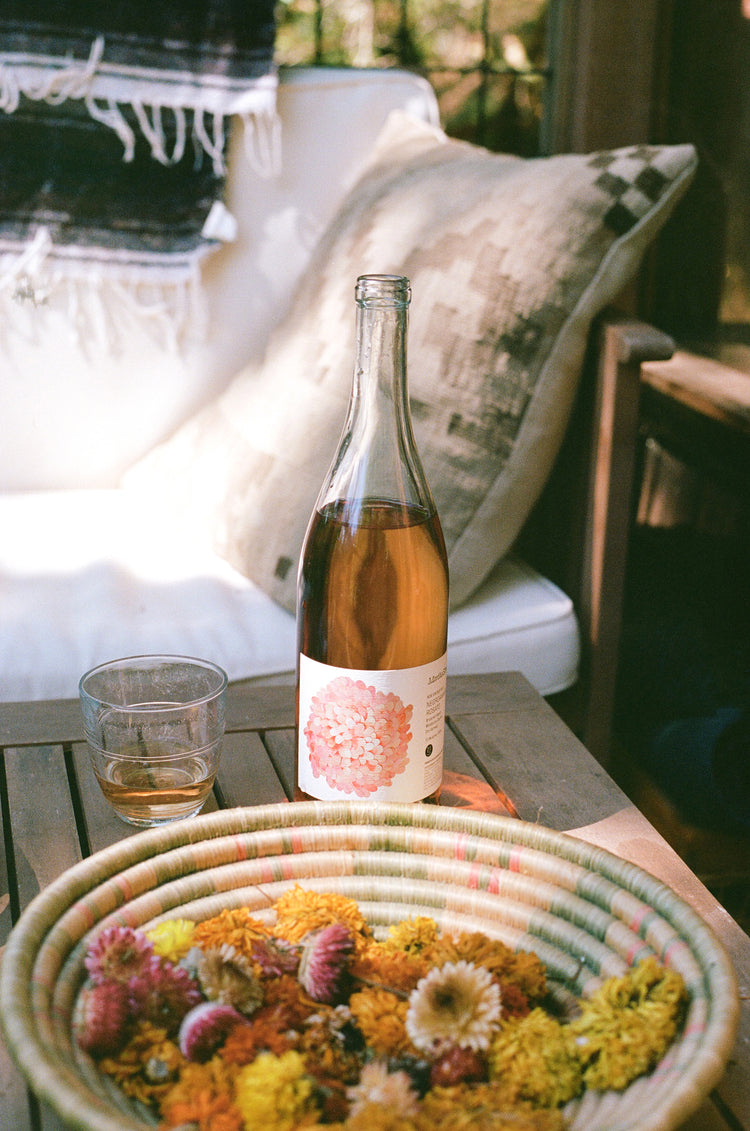 A cozy setting featuring a bottle of wine, a glass, and a basket of colorful dried flowers on a wooden table.