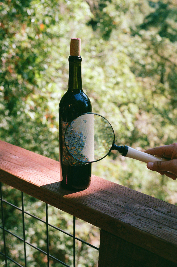 A wine bottle on a wooden railing, with a magnifying glass focusing on its label. Lush greenery is blurred in the background.