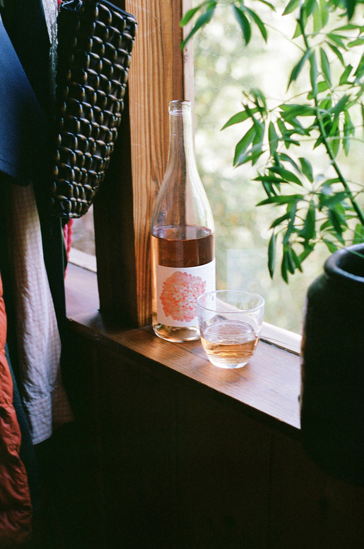 A bottle of drink with a floral label and a glass sit on a wooden windowsill, surrounded by greenery and hanging items.