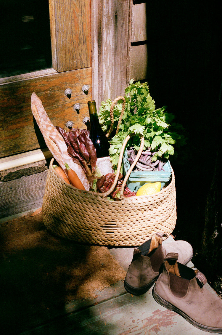 A woven basket filled with fresh vegetables, herbs, and a bottle of wine, placed near a wooden door and a pair of shoes.