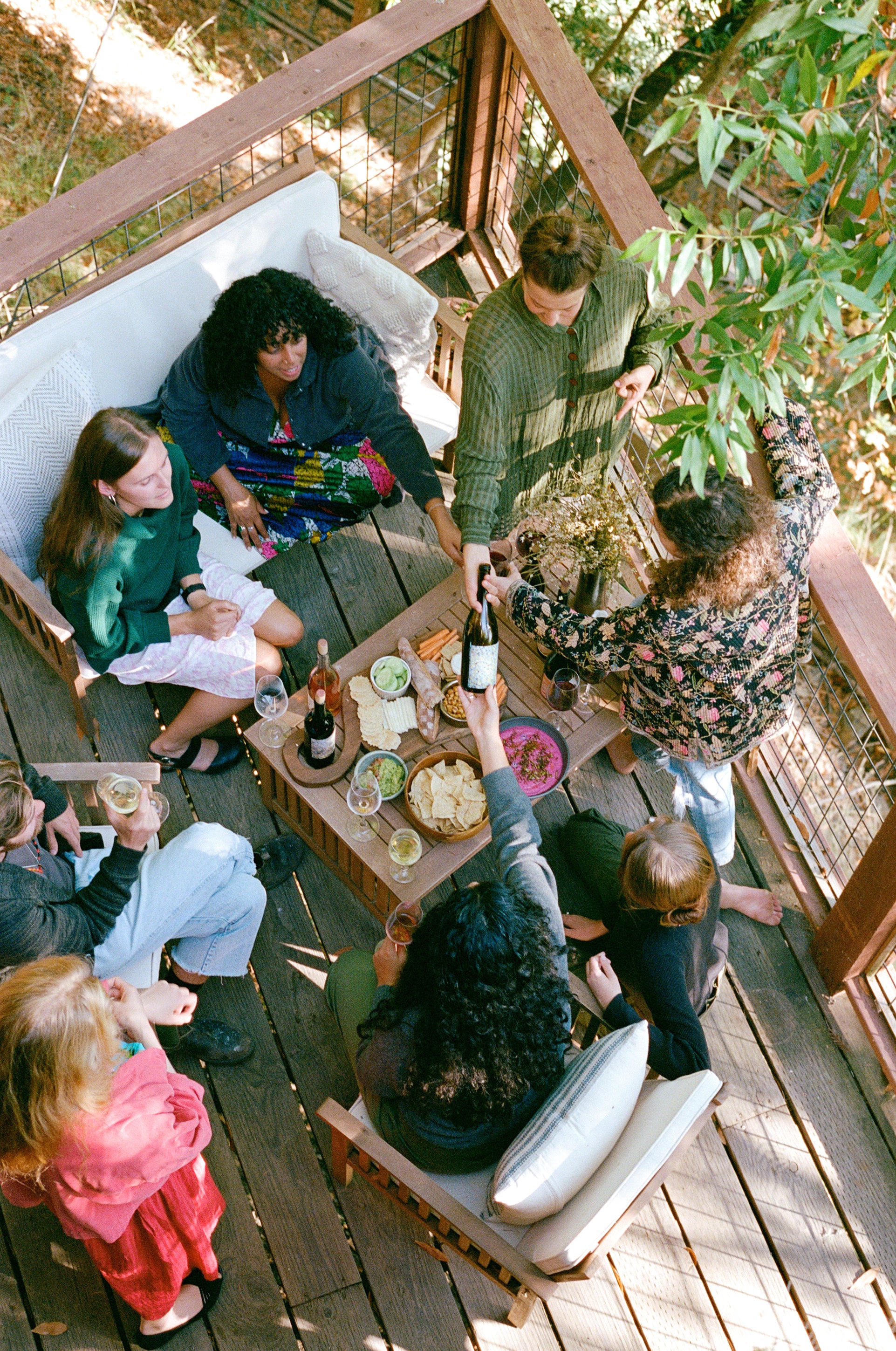 A group of people enjoying drinks and snacks on a wooden deck, surrounded by greenery.