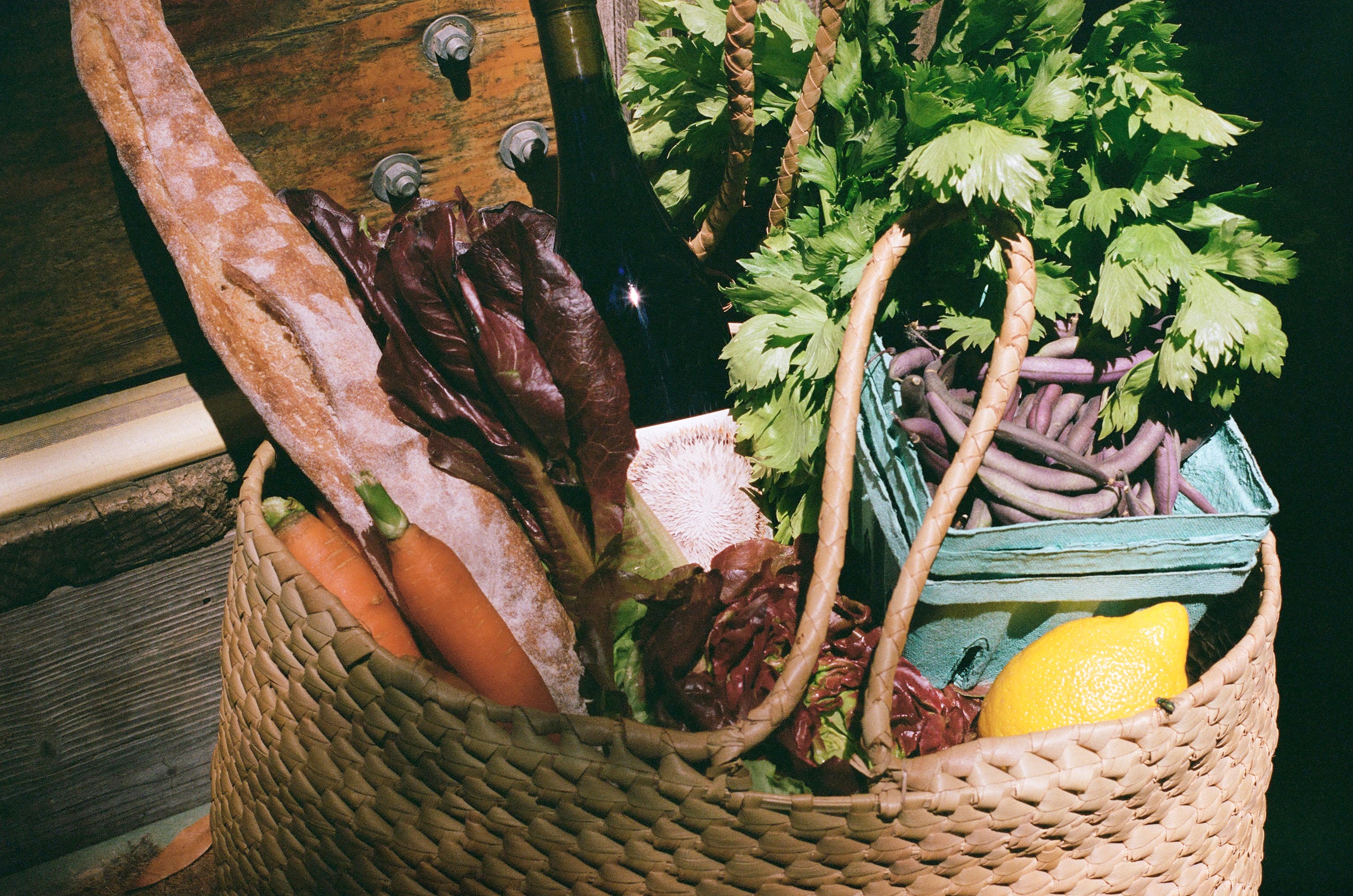 A woven basket filled with fresh produce including a baguette, carrots, red lettuce, celery, a lemon, and beans. A wine bottle is tucked inside, conveying a rustic and wholesome vibe.