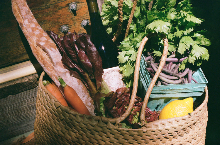 A woven basket filled with fresh produce including a baguette, carrots, red lettuce, celery, a lemon, and beans. A wine bottle is tucked inside, conveying a rustic and wholesome vibe.