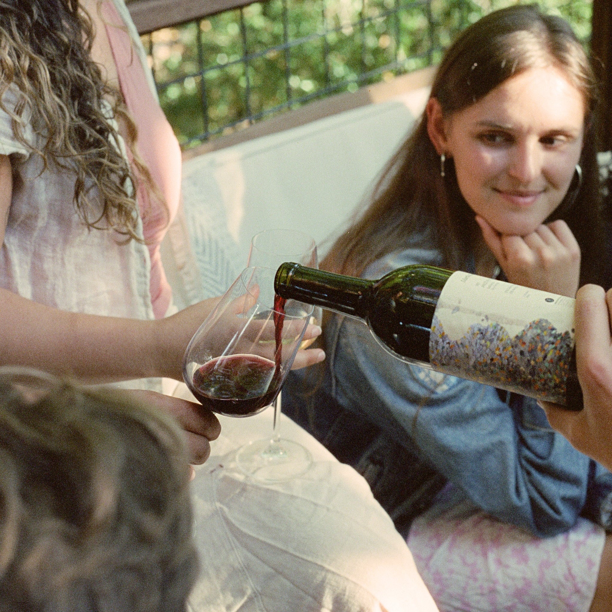 A group of friends enjoying wine outdoors, with one person pouring red wine into a glass while others watch and smile.