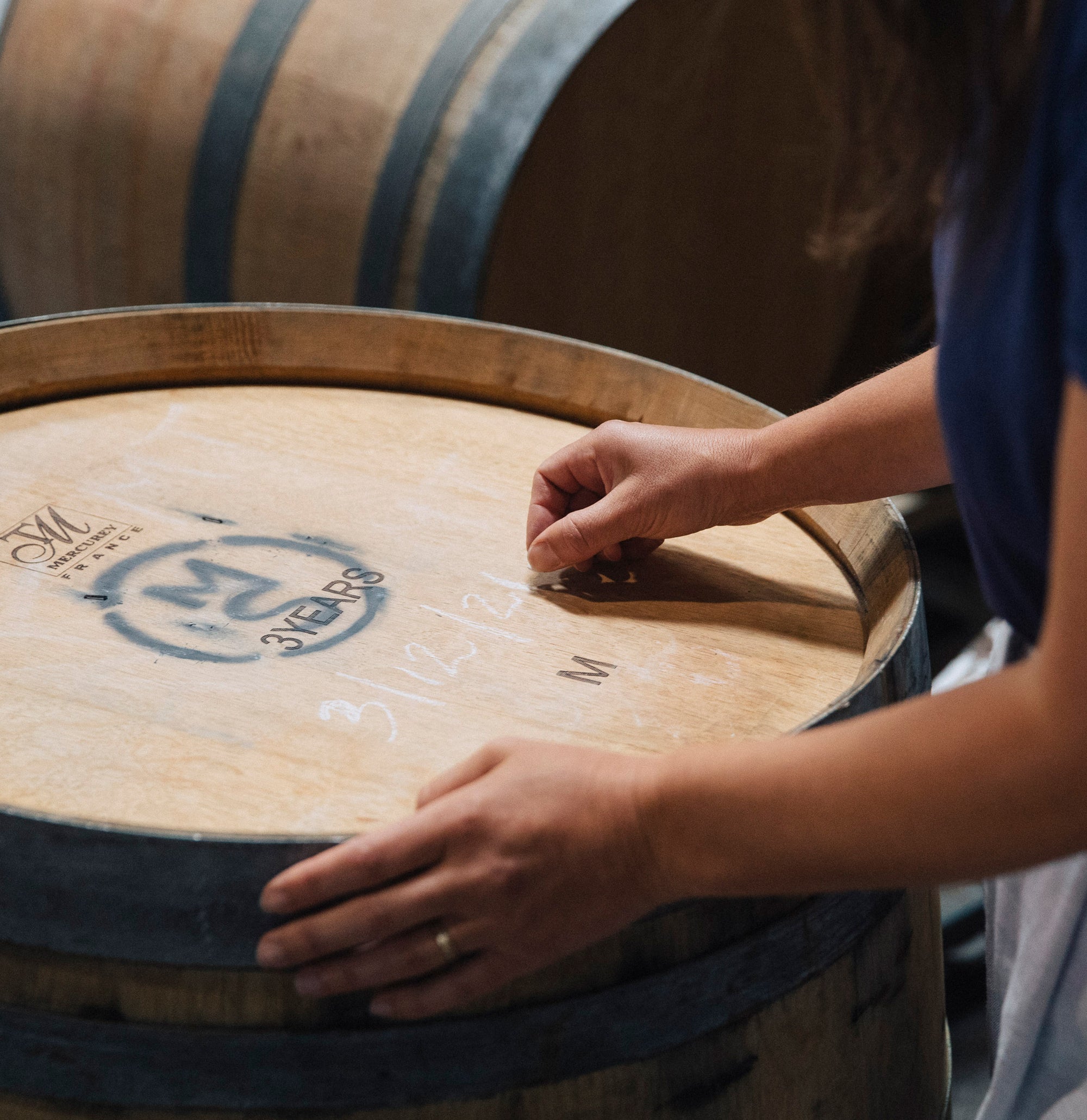 A person writes on the lid of a wooden barrel labeled "3 Years" in chalk. The setting is rustic, conveying a sense of tradition and craftsmanship.