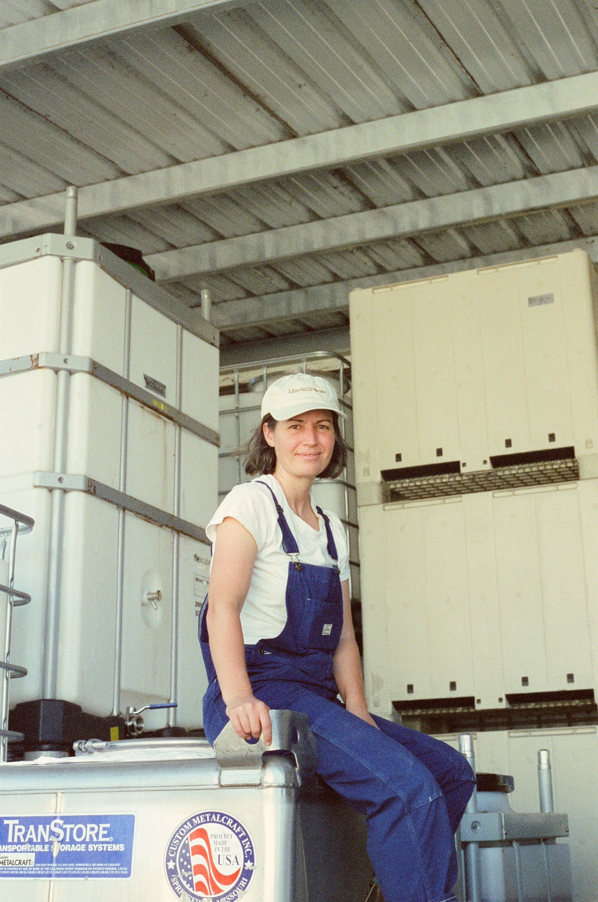 A person in blue overalls and a white cap sits on a metal container in an industrial setting, surrounded by storage units. They are smiling confidently.