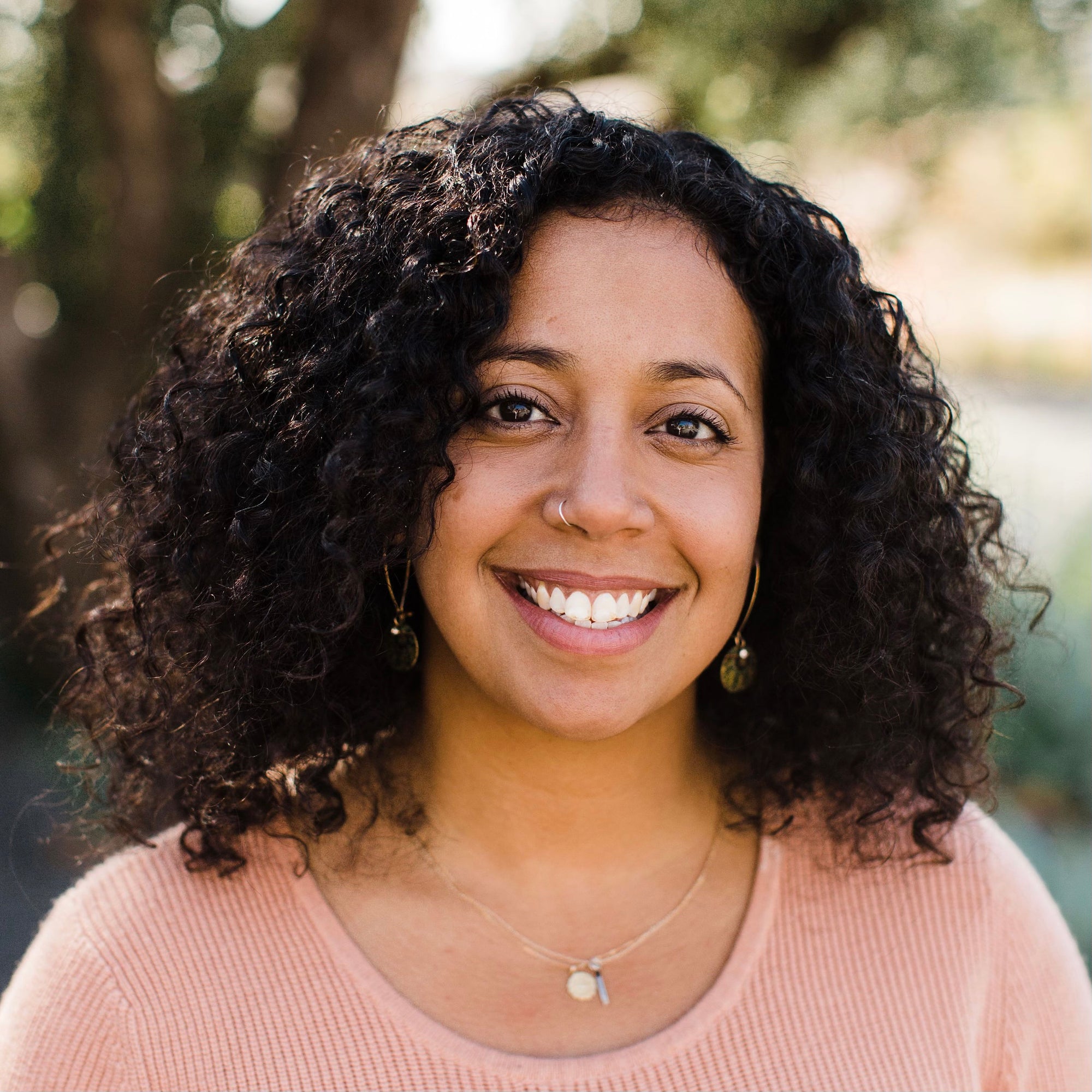 Smiling woman with curly hair and a nose ring, wearing a peach sweater. She stands outdoors with a blurred, sunny background, conveying warmth and friendliness.