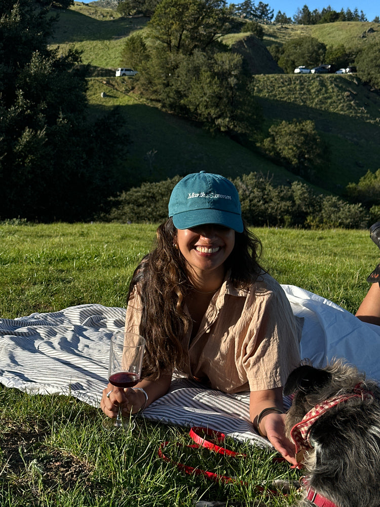 A woman wearing a teal Martha Stoumen hat lays in the grass with ehr dog and a glass of wine.