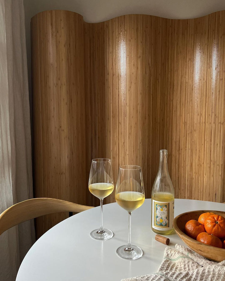 A cozy table setting with two glasses of yellow drink, a bottle, and a bowl of oranges against a bamboo backdrop.
