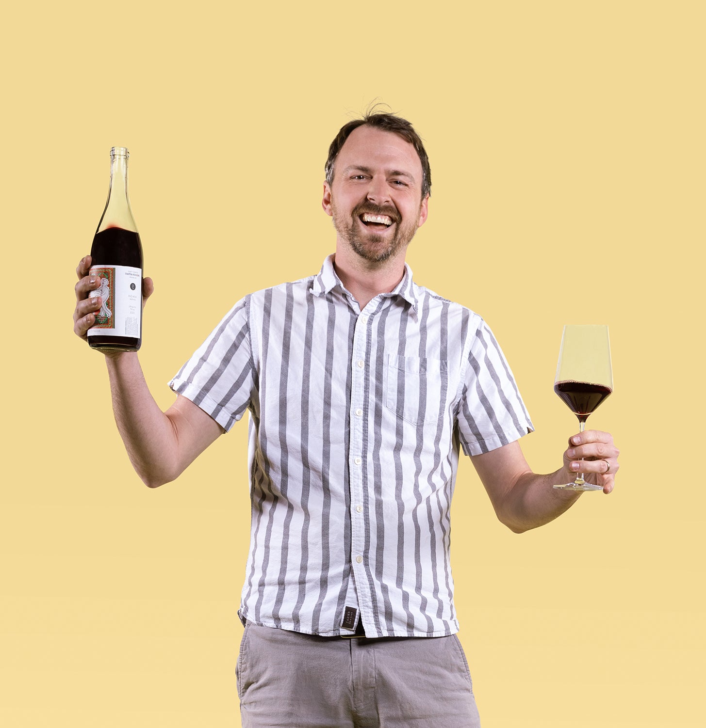 Smiling man holding a bottle of red wine and a glass, wearing a striped shirt against a yellow background. The mood is joyful and celebratory.