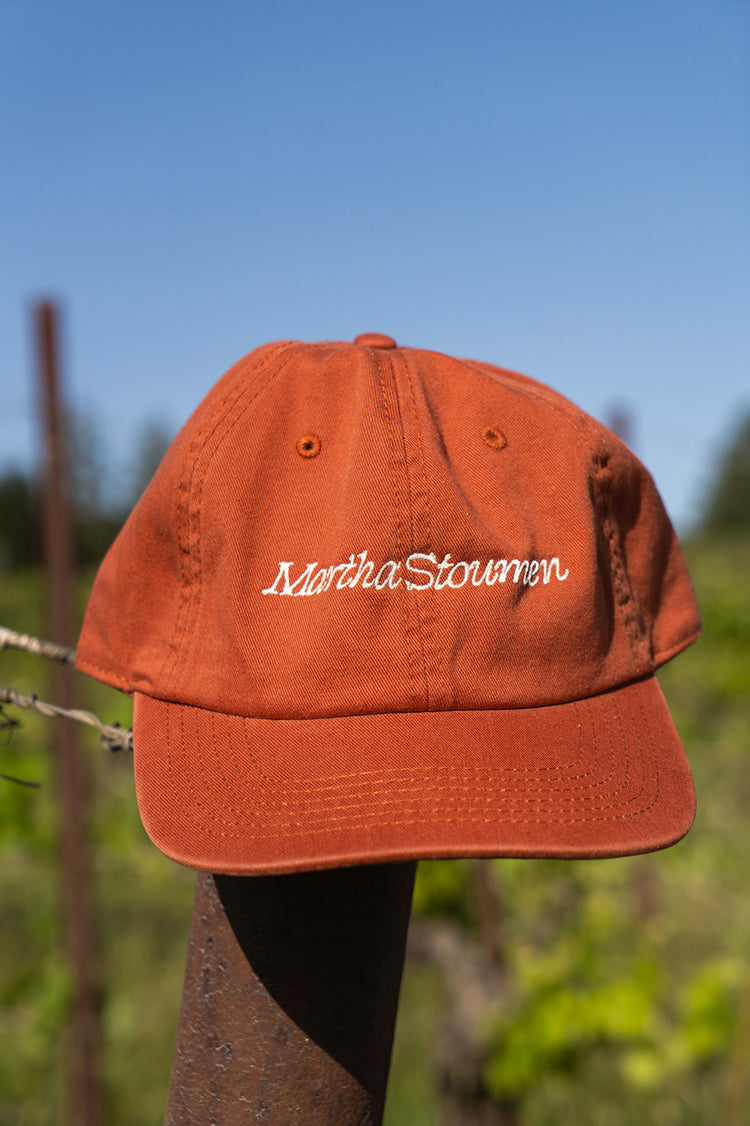 An orange cap with white embroidered text reading "Martha Stoumen," resting on a metal post in a vineyard setting.