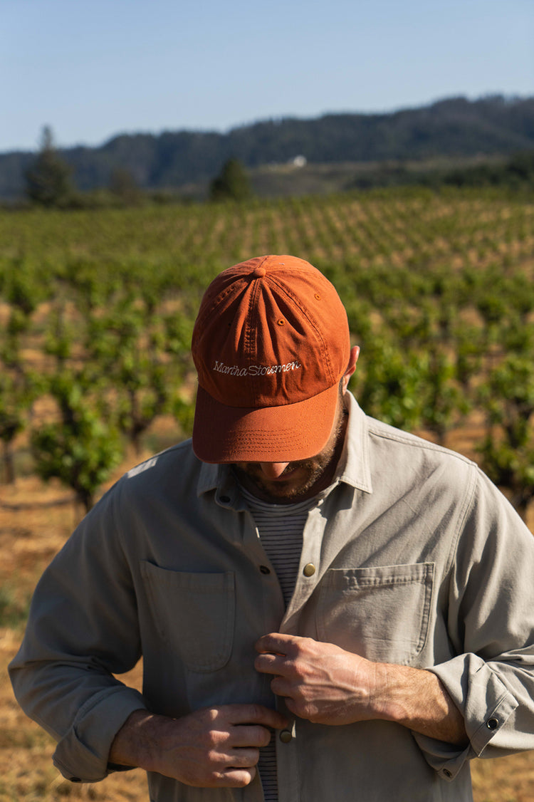 A man in a brown cap and gray shirt adjusts his clothing in a vineyard, with rows of grapevines and hills in the background.