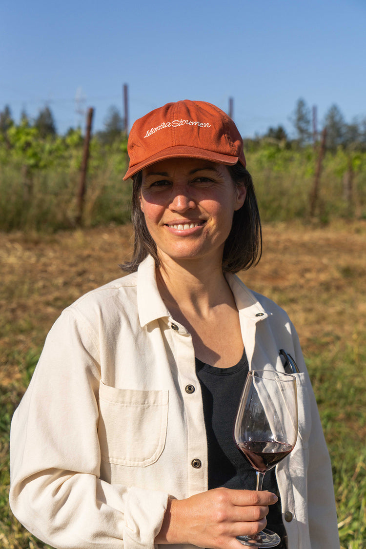 A woman in a vineyard smiles warmly, wearing a rust-colored cap and beige shirt, holding a glass of red wine. The mood is relaxed and joyful.