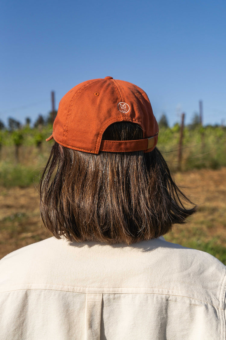 A person with shoulder-length hair wearing an orange cap stands in a vineyard under a clear blue sky.