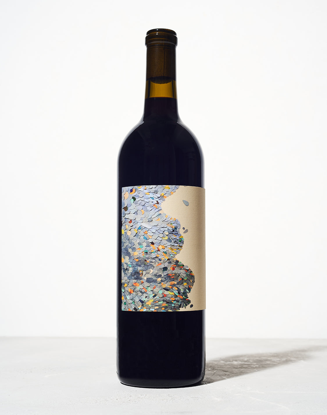A wine bottle with a dark glass is centered against a white background. The label features a colorful abstract pattern of overlapping leaf-like shapes.