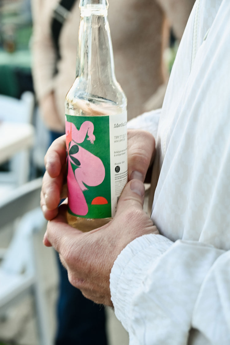 A person holds a glass bottle with a colorful label featuring a pink figure and green background.