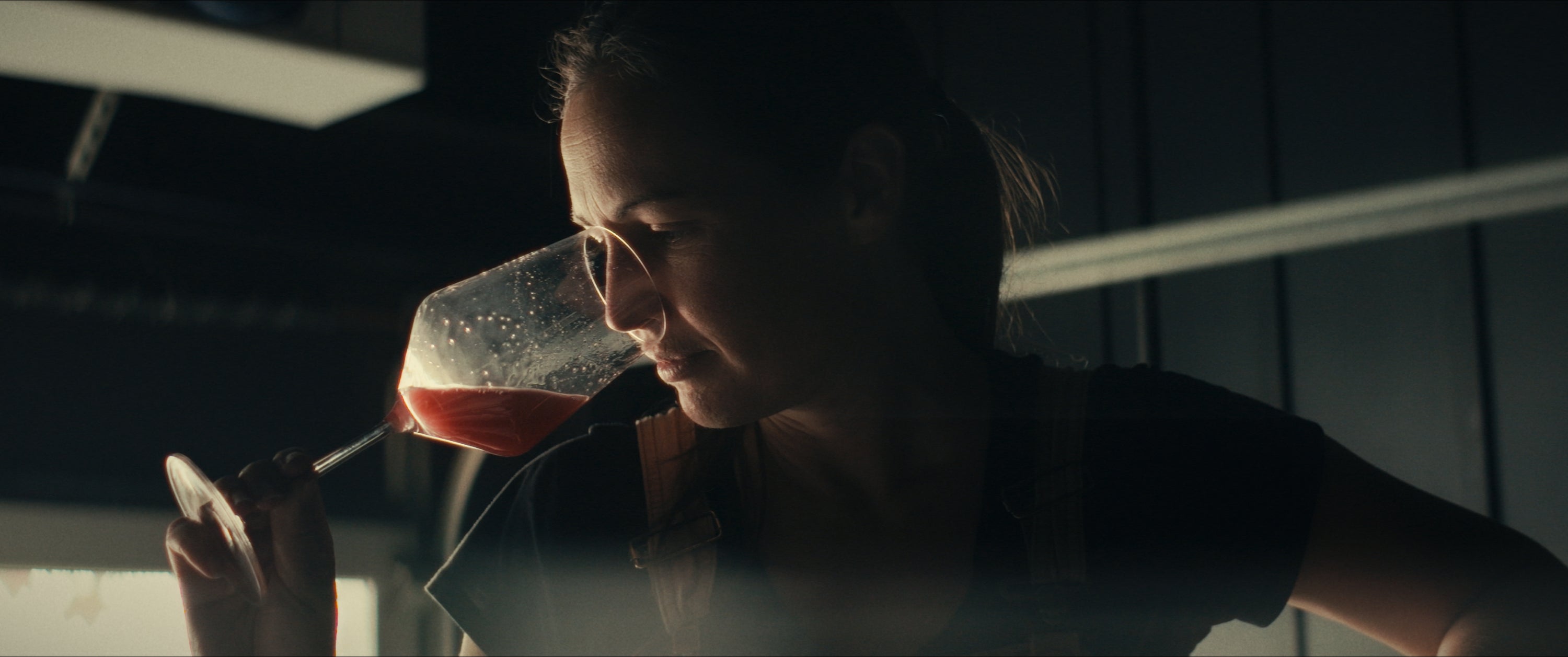 A woman examines a glass of red liquid, holding it close to her face in a dimly lit setting.