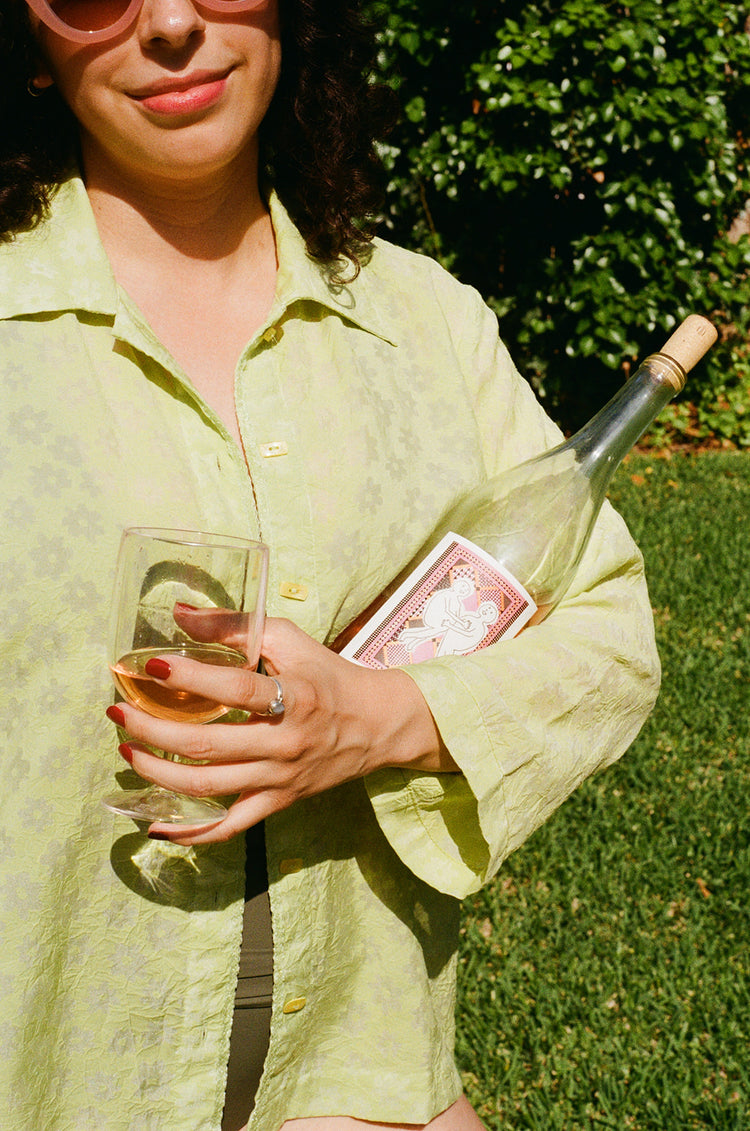 A person in a light green shirt holds a glass of wine and a bottle with a playful label, surrounded by greenery.