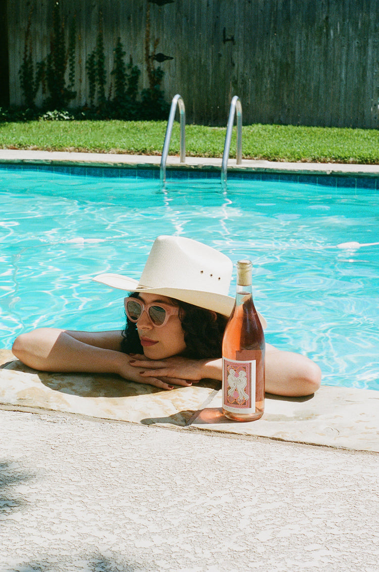 A person with curly hair, wearing a wide-brimmed hat and sunglasses, relaxes by a pool with a bottle of rosé wine.