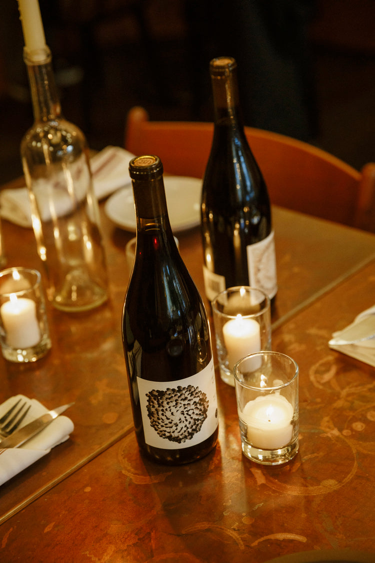 Two bottles of wine sit on a copper table with votive candles. The background bottle is blurred slightly, and the bottle in the foreground features a black rosetta style label.
