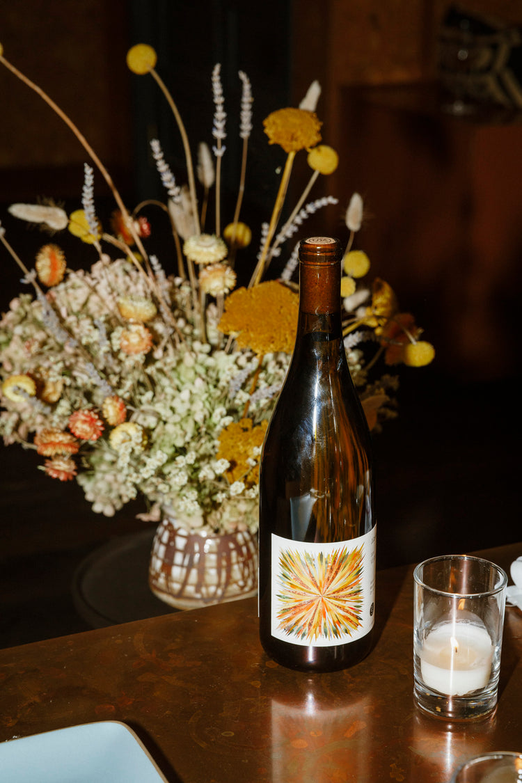 A bottle of Martha Stoumen Vermentino wine — a brown glass bottle with a vibrant yellow and orange starburst label— sits in front of a dried floral arrangement on a copper table top.