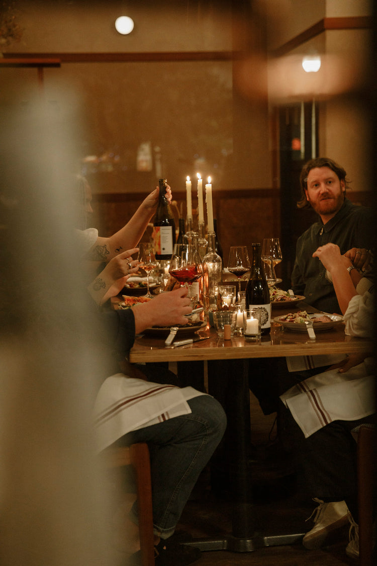 This image features a dinner party, captured from the outside and through some cafe curtains abstracting the foreground. There is a bottle of wine on the table, a bottle being passed acrossed the table, and men and women all intermingled at dinner.