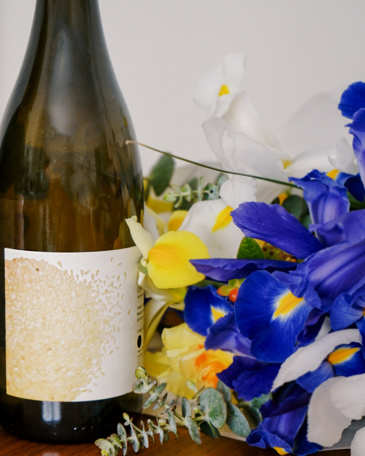 A bottle with an abstract label sits next to vibrant blue, yellow, and white irises. The arrangement evokes a fresh and elegant atmosphere.