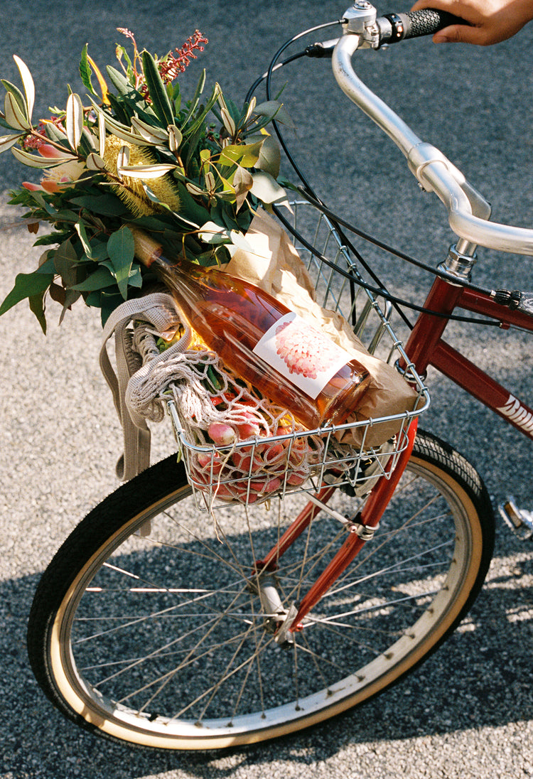 A bicycle with a wire basket holds a bouquet of flowers, a bottle of rosé wine, and colorful fruits.