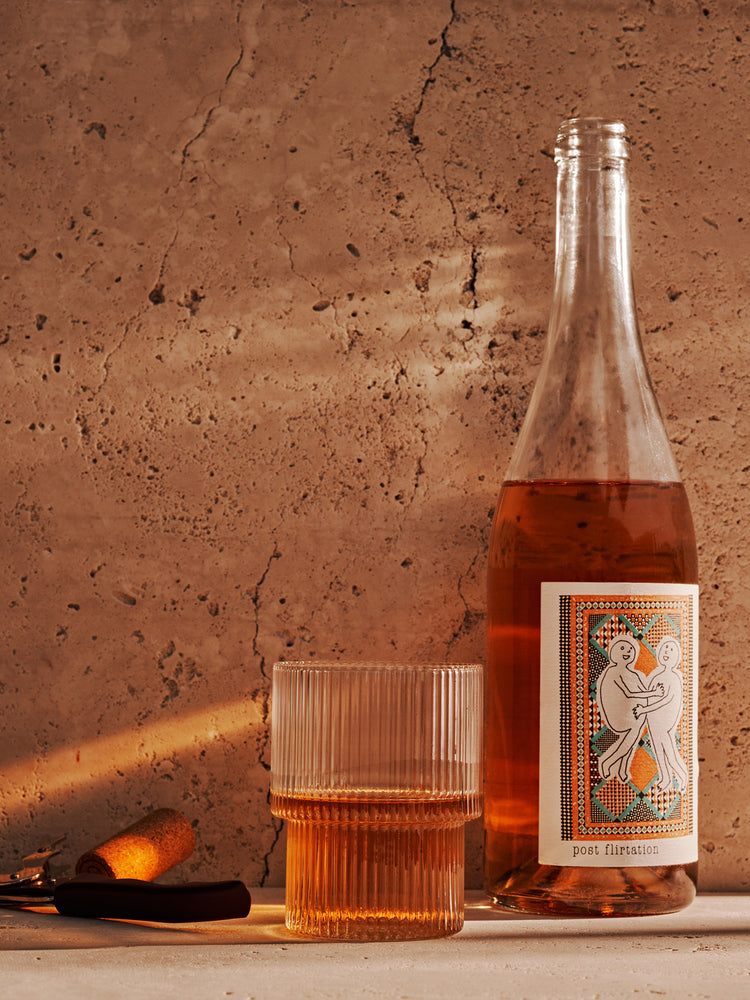 A bottle of rosé wine with a decorative label sits next to a ribbed glass filled with wine, on a textured surface.
