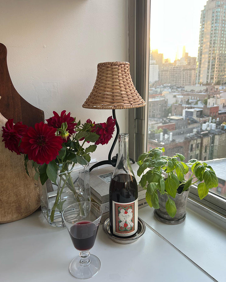 A cozy table scene featuring a bottle of wine, a glass, vibrant red flowers, and a small potted plant, with a city view in the background.