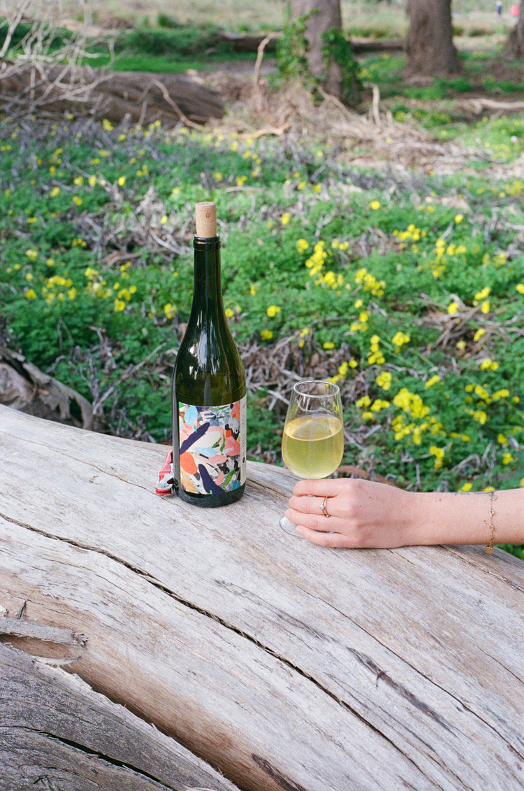 Bottle of wine with a colorful label and a glass of wine on a wooden surface with a natural background.