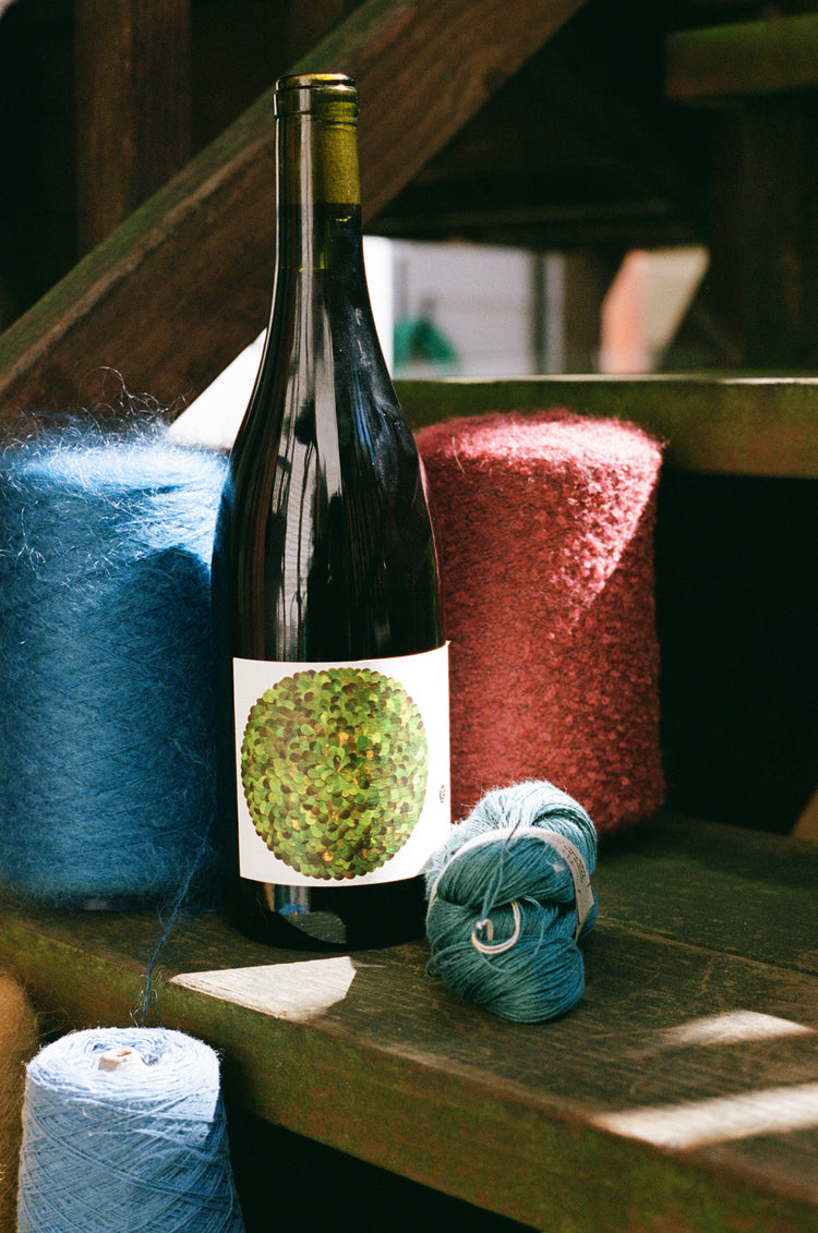 Bottle of wine with a green label next to skeins of yarn on a wooden surface.
