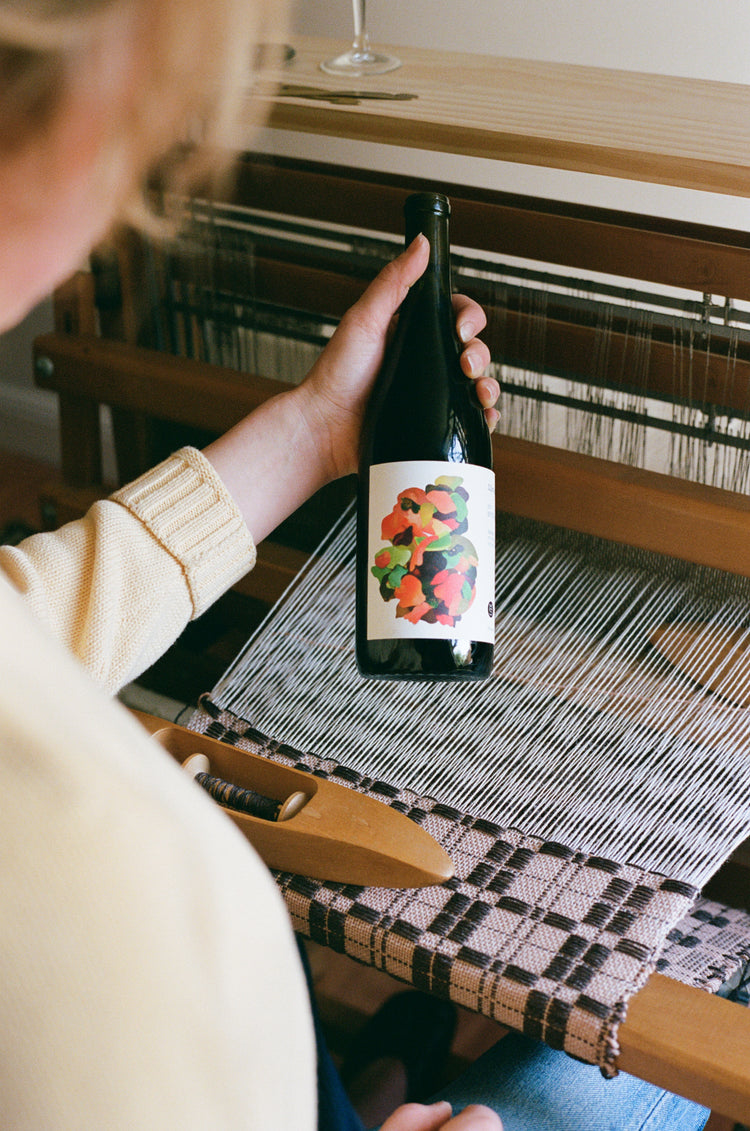 A person holding a dark glass bottle with a colorful label in front of a wooden loom.
