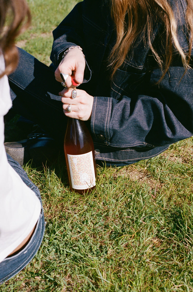 A woman sitting in the grass in all denim opening a bottle of wine with a confetti label