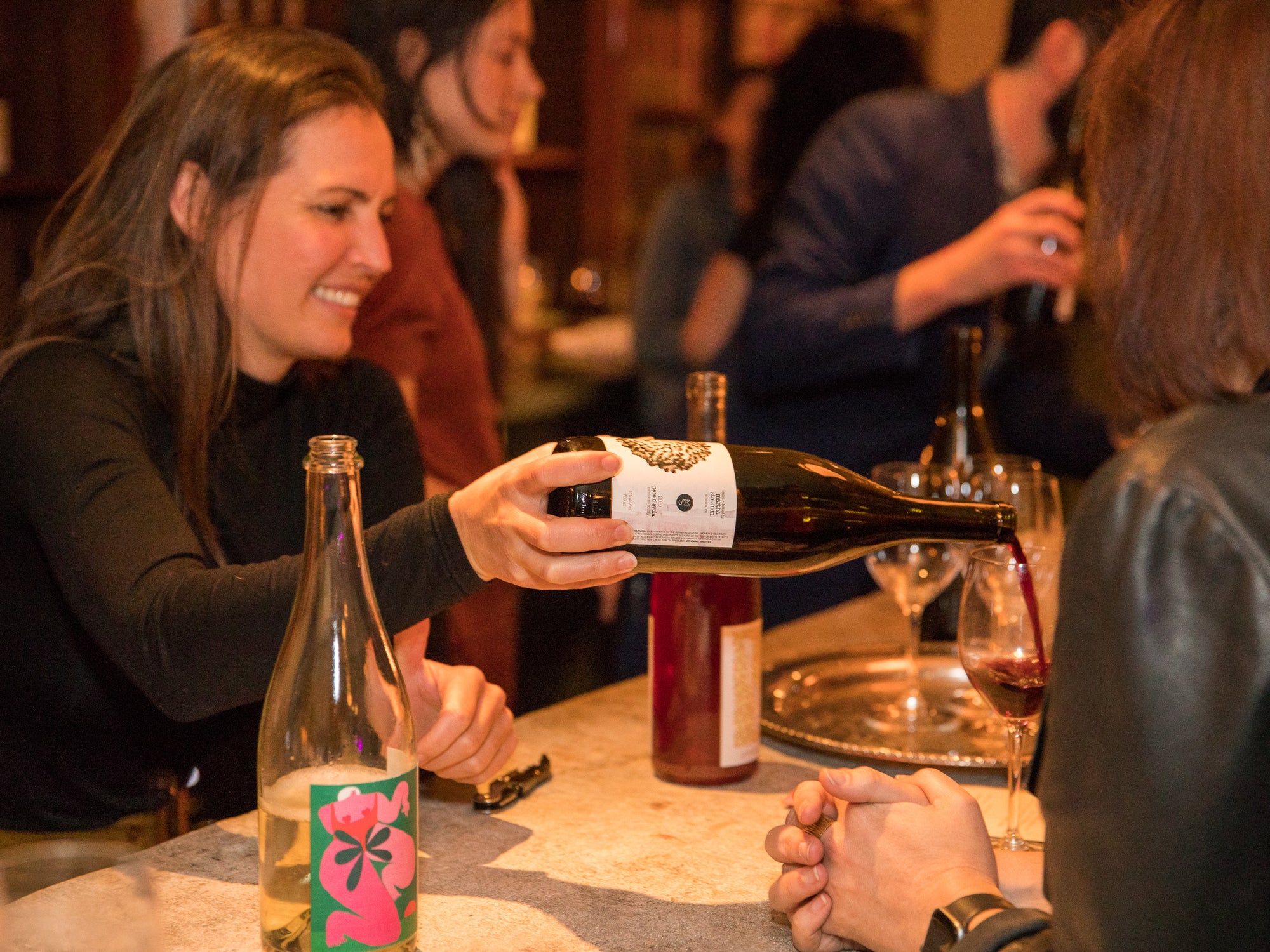 A woman pours wine from a bottle into a glass at a social gathering, with other people and bottles in the background.