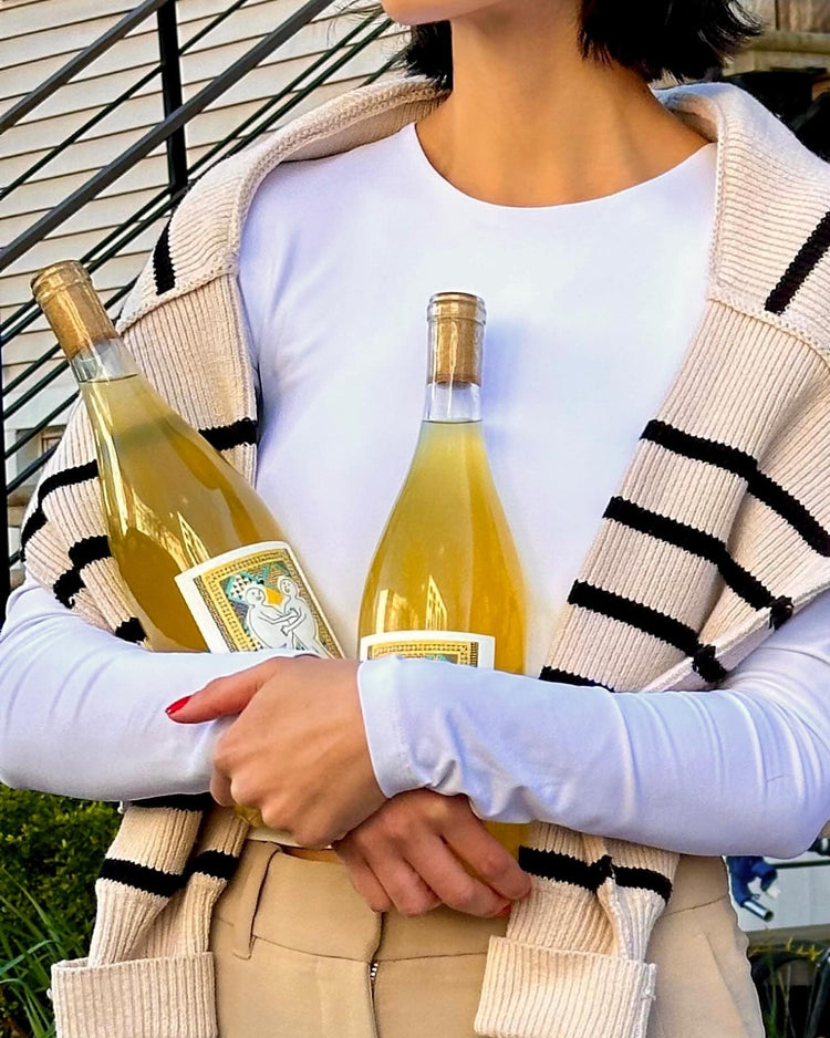 A person holds two bottles of yellow liquid, wearing a white top and a striped cardigan, with a staircase in the background.