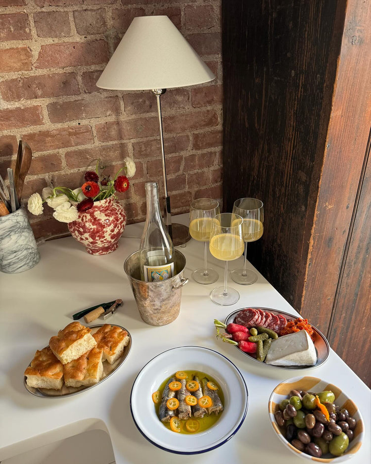 A cozy table setting featuring a lamp, flowers, drinks, and an assortment of food including bread, olives, and fish.