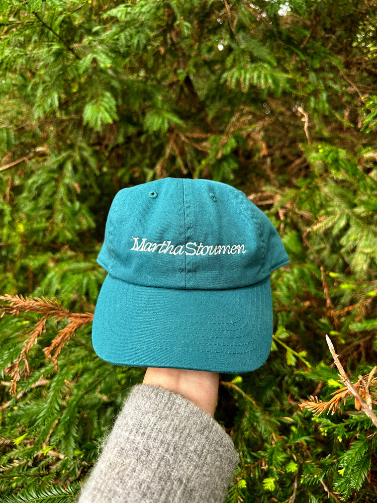 A teal cap with "martha stoumen" embroidered on the front, being held in front of a redwood tree