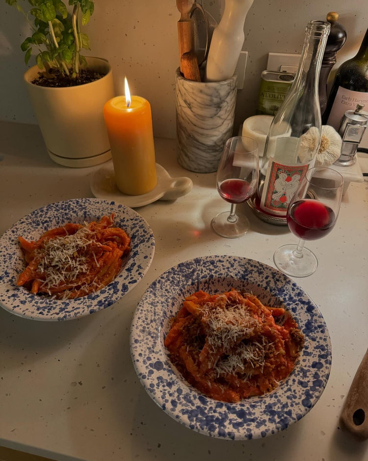 Two plates of pasta topped with cheese, a lit candle, wine glasses, and a bottle of wine on a kitchen counter.