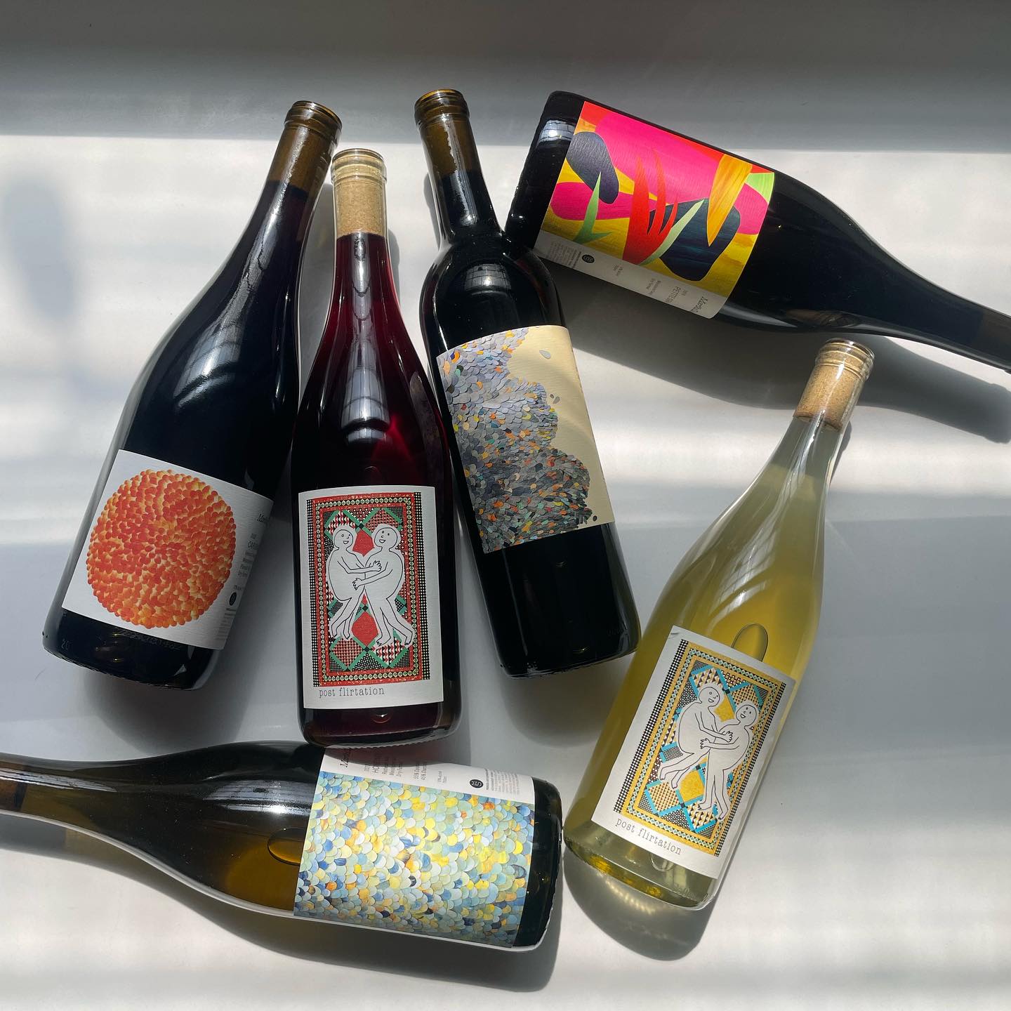 Six wine bottles with colorful, artistic labels are arranged on a sunlit surface. Labels feature abstract designs and vibrant colors, evoking a playful tone.