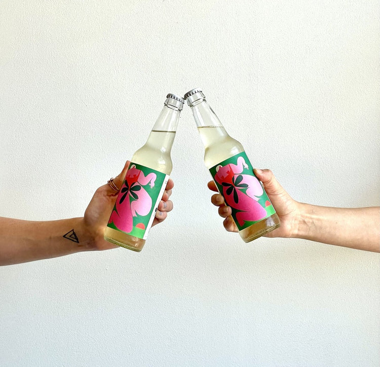 Two hands clink colorful beverage bottles against a plain background. The bottles feature vibrant floral designs.