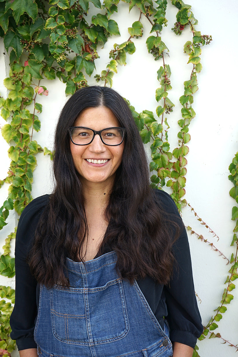 A smiling woman with long dark hair and glasses stands in front of a white wall covered in green ivy. She wears a denim apron over a black shirt.
