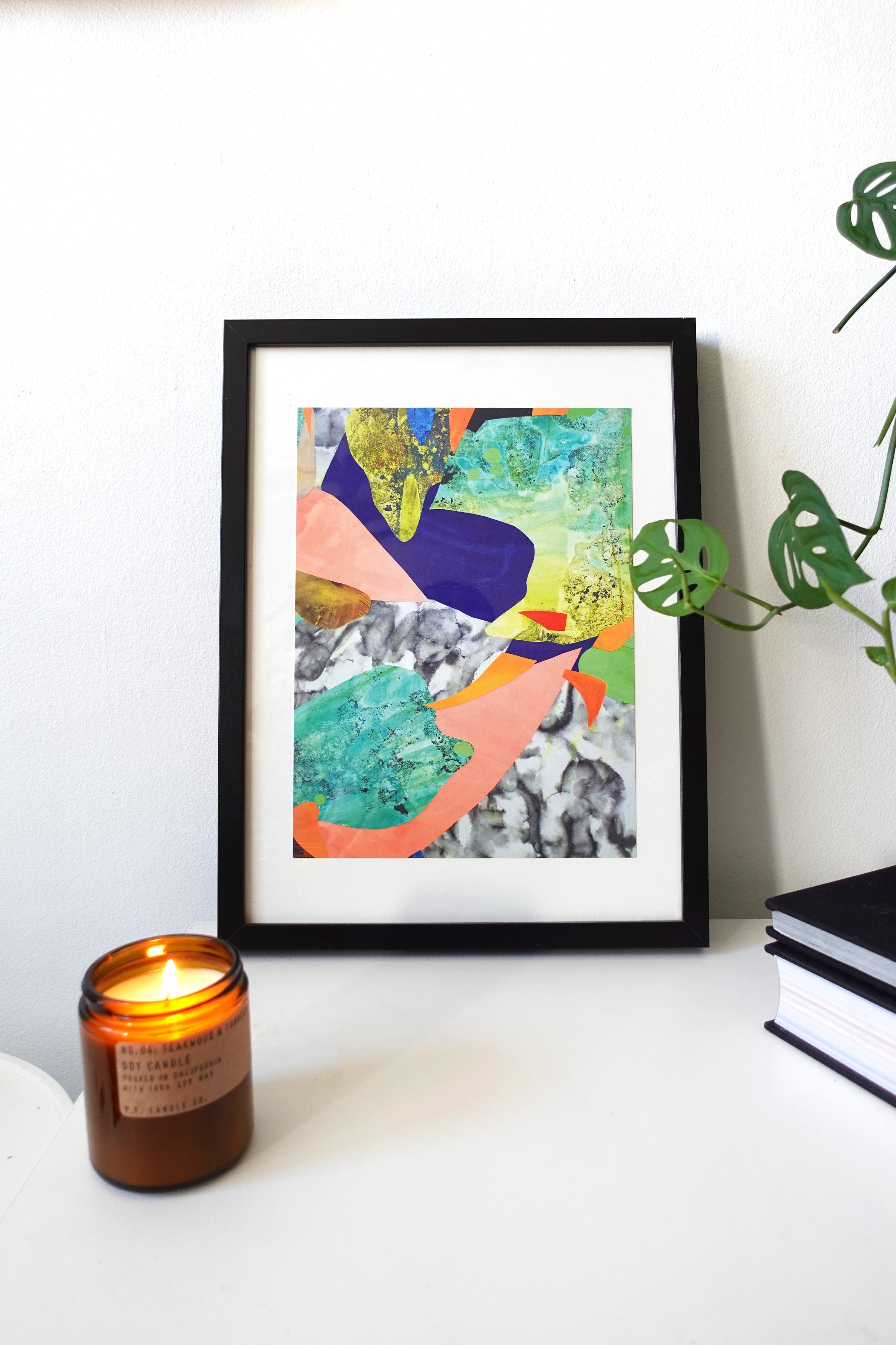 Framed abstract art with vibrant colors like blue, green, and orange. A lit brown candle and a small plant add warmth and serenity to the scene.