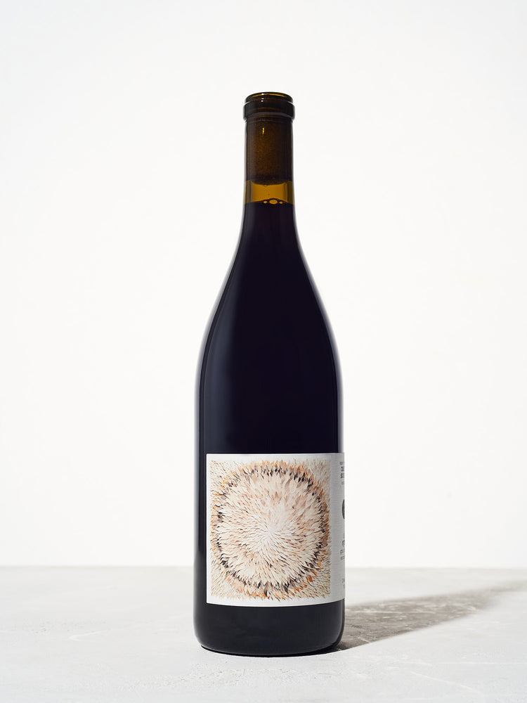 A wine bottle with a dark glass body and a minimalist label featuring an abstract pattern of beige and brown brushstrokes, standing on a neutral surface.