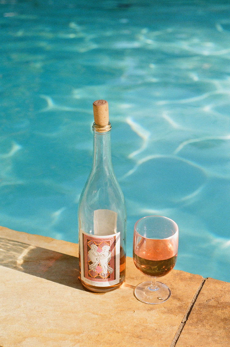 A wine bottle and glass of rosé sit on a stone edge by a clear, shimmering pool under sunlight, evoking a relaxed and summery atmosphere.