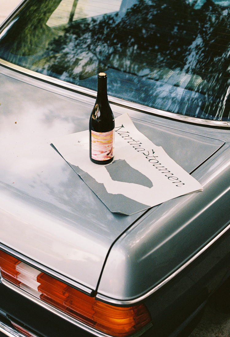 A wine bottle sits atop a gray car trunk, surrounded by a printed paper with "Martha" visible. The scene has a relaxed, casual vibe.